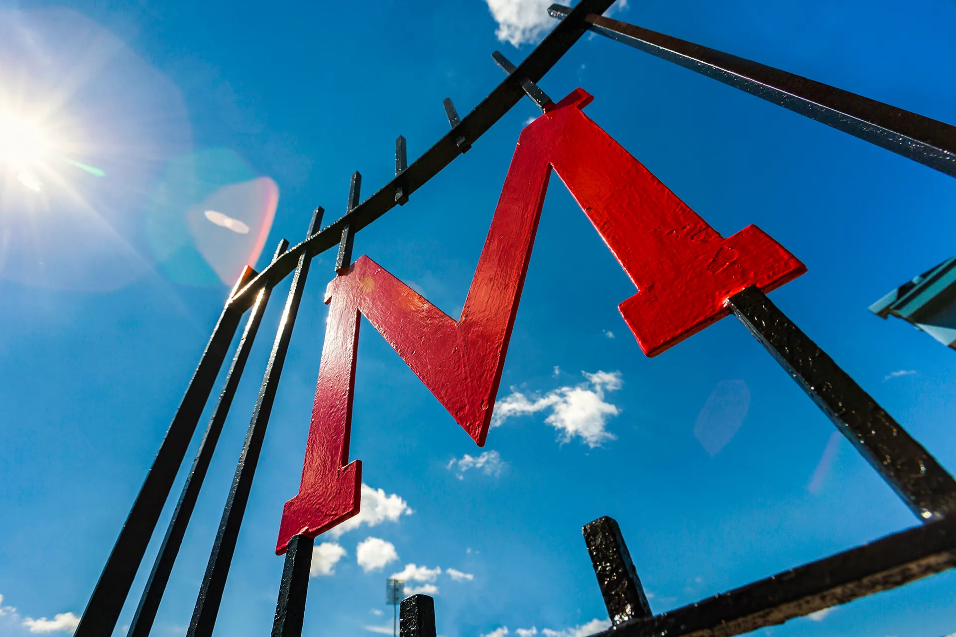 A bright, sunny day with a clear blue sky, where sunlight creates a lens flare effect. A red metallic "M" is mounted on black metal gate or fence.