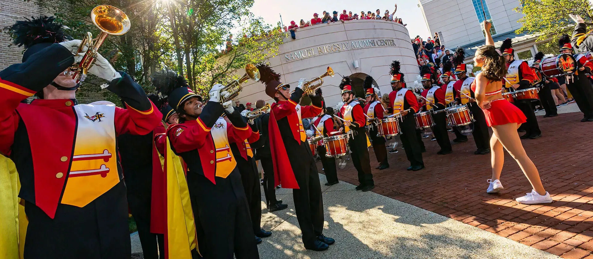 Marching band playing - trumpet in foreground, other brass players, drummers. A cheerleader in front. People watching in the background from on top of a modern building: Alumni Center
