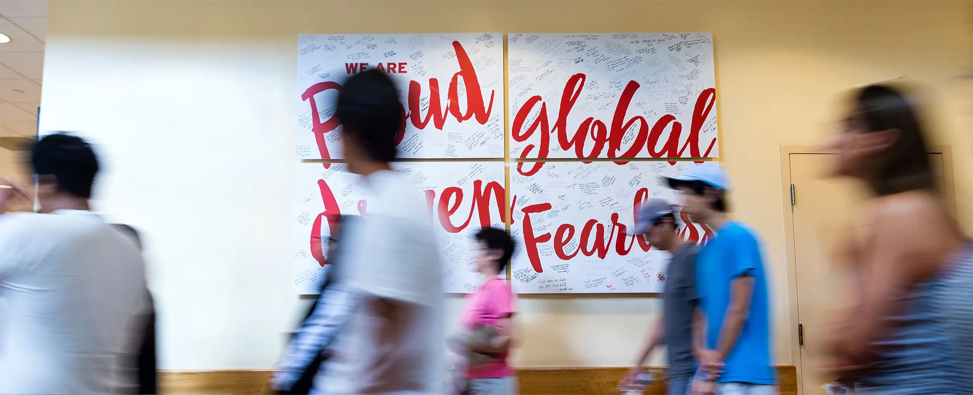 motion blur of people walking in front of signs:"We are: Proud, global, driven, Fearless"