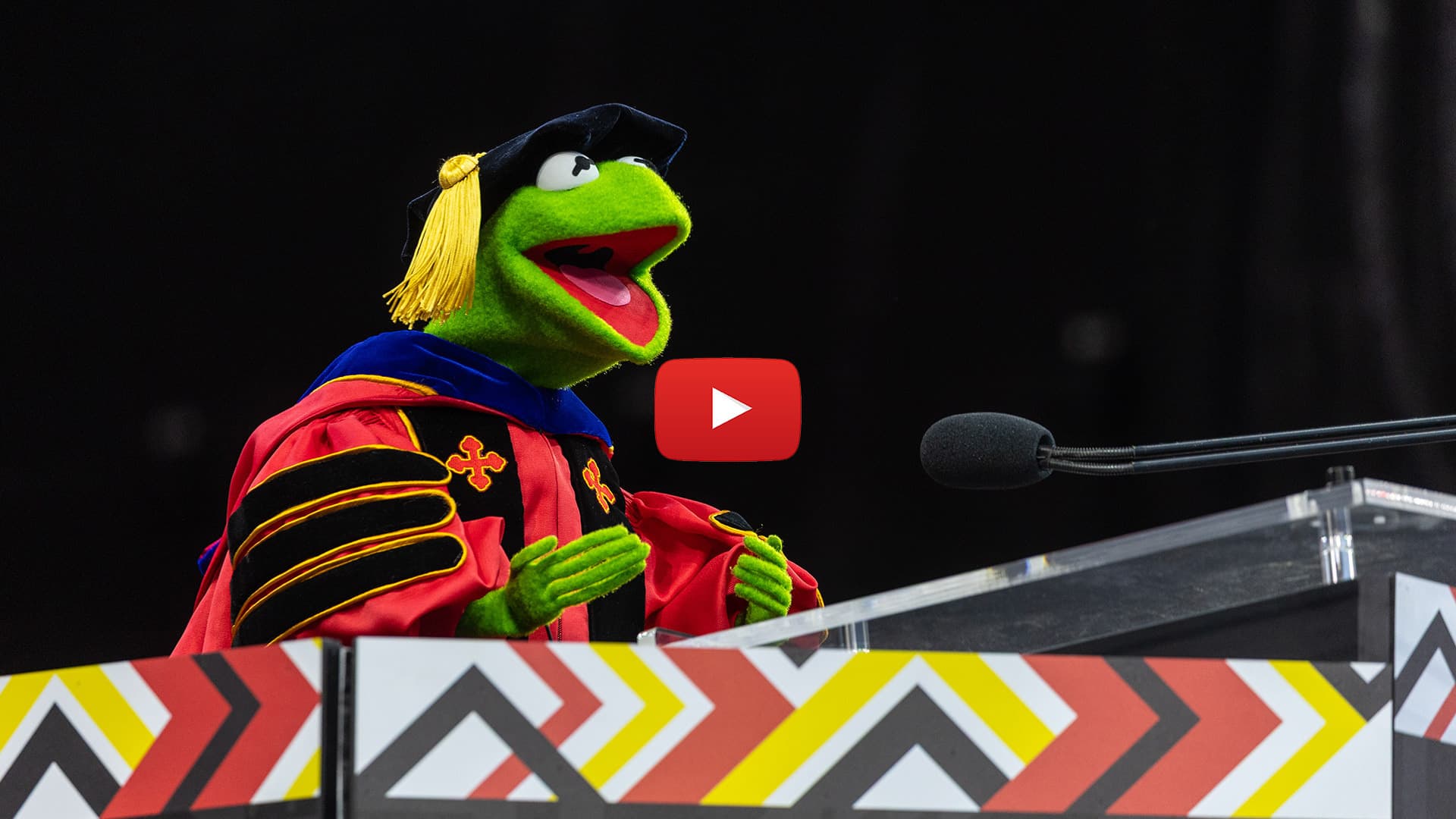 Kermit the Frog wearing regalia and speaking at UMD commencement, with red video play button