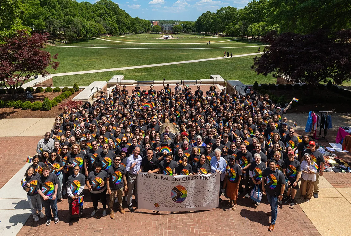 Large group of students and some faculty. Most wearing the same t-shirt - rainbow turtle shell with chevron. In the front, students hold a sign: "Inaugural Big Queer Photo, May 2, 2024, LGBTQ+ Equity Center". Sign has many signatures