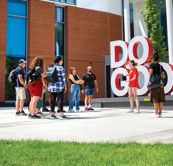Prospective students on a campus tour, outside a modern building with "Do Good" sculpture in front.