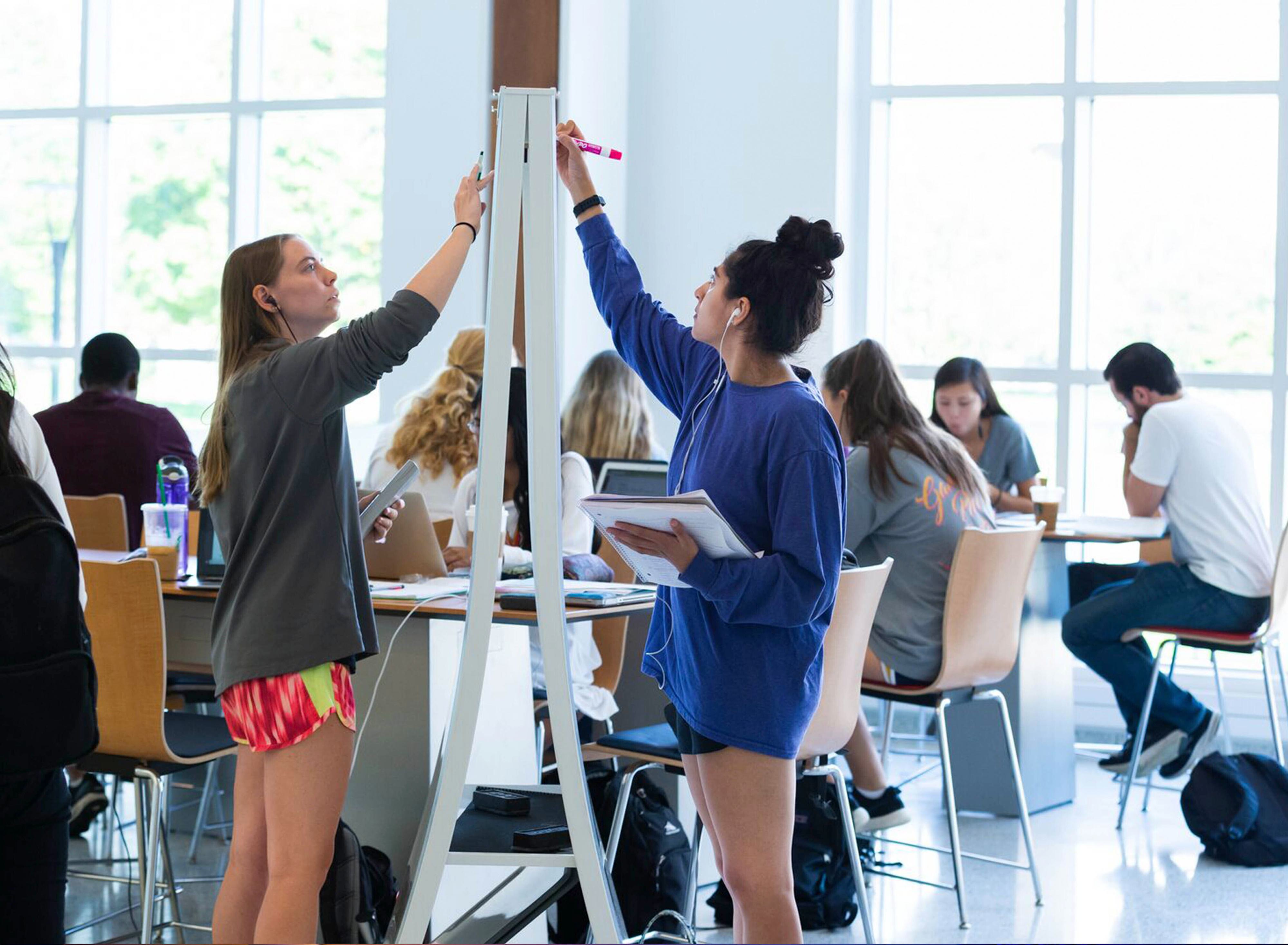 Two students stand on opposite sides of a dry erase board, each reaching up to write on the side facing them.