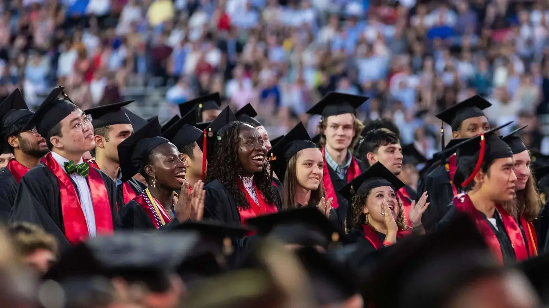 group of university graduates in black caps and gowns with red stoles stand and cheer during a commencement ceremony