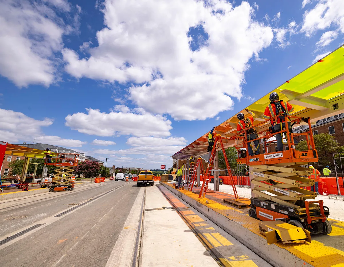 Construction on a rail line. Track is visible, construction workers on lifts, vehicles in background. Train stop awning is being worked on.