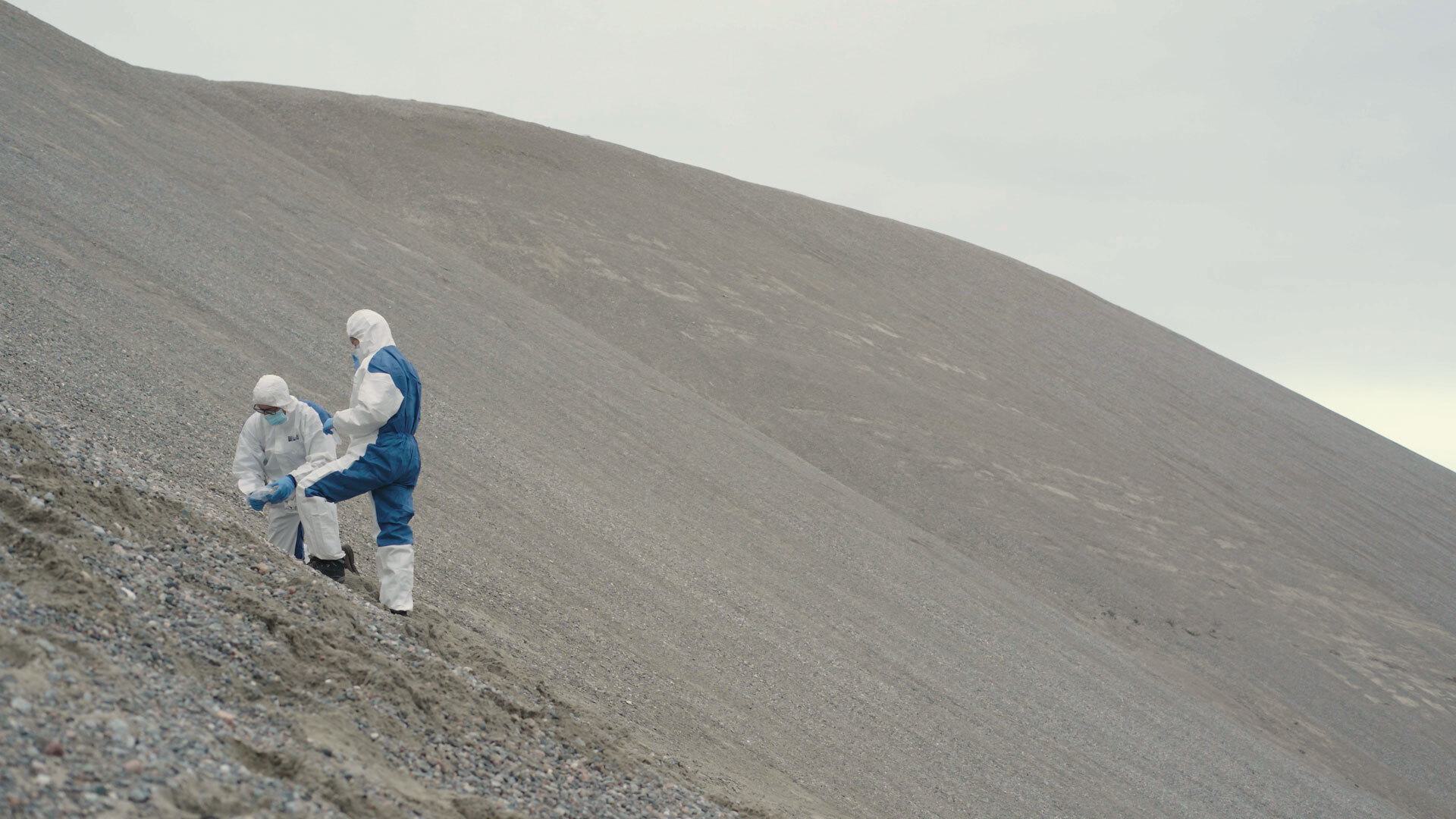 two researchers wearing hazmat suits sample sediment on hill
