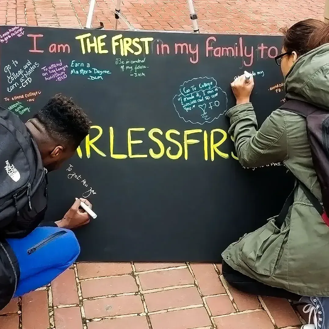 2 students outside writing on a chalkboard that's titled "First in my family to…" Answers include "go to college", "get a master's degree"