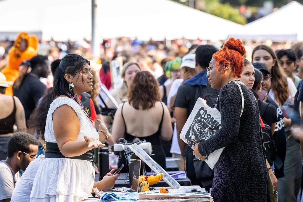 Two students talk to one another across an event table with many more students and tents in the background