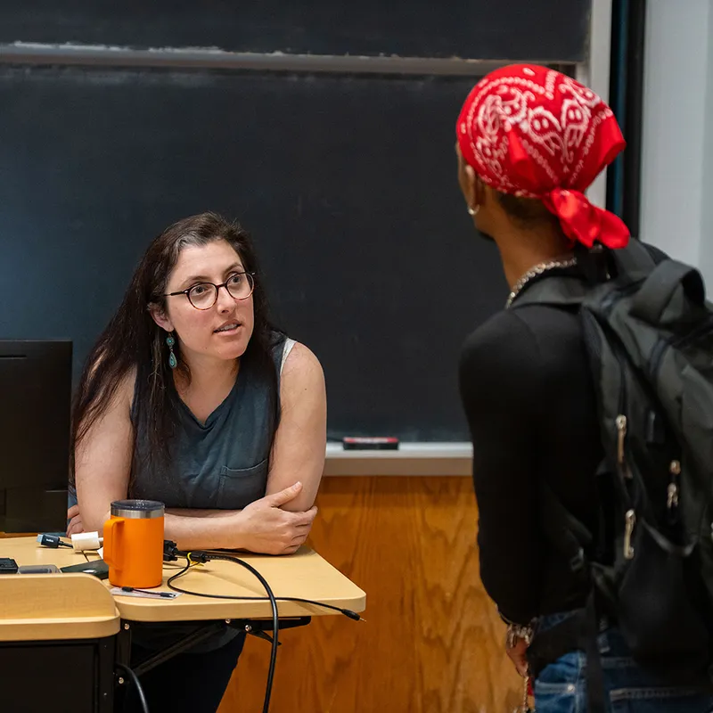 This image depicts an interaction between a professor and a student. The professor, seated behind a desk in front of a blackboard, is engaged in conversation with the standing student, who has a red bandana tied around their head.