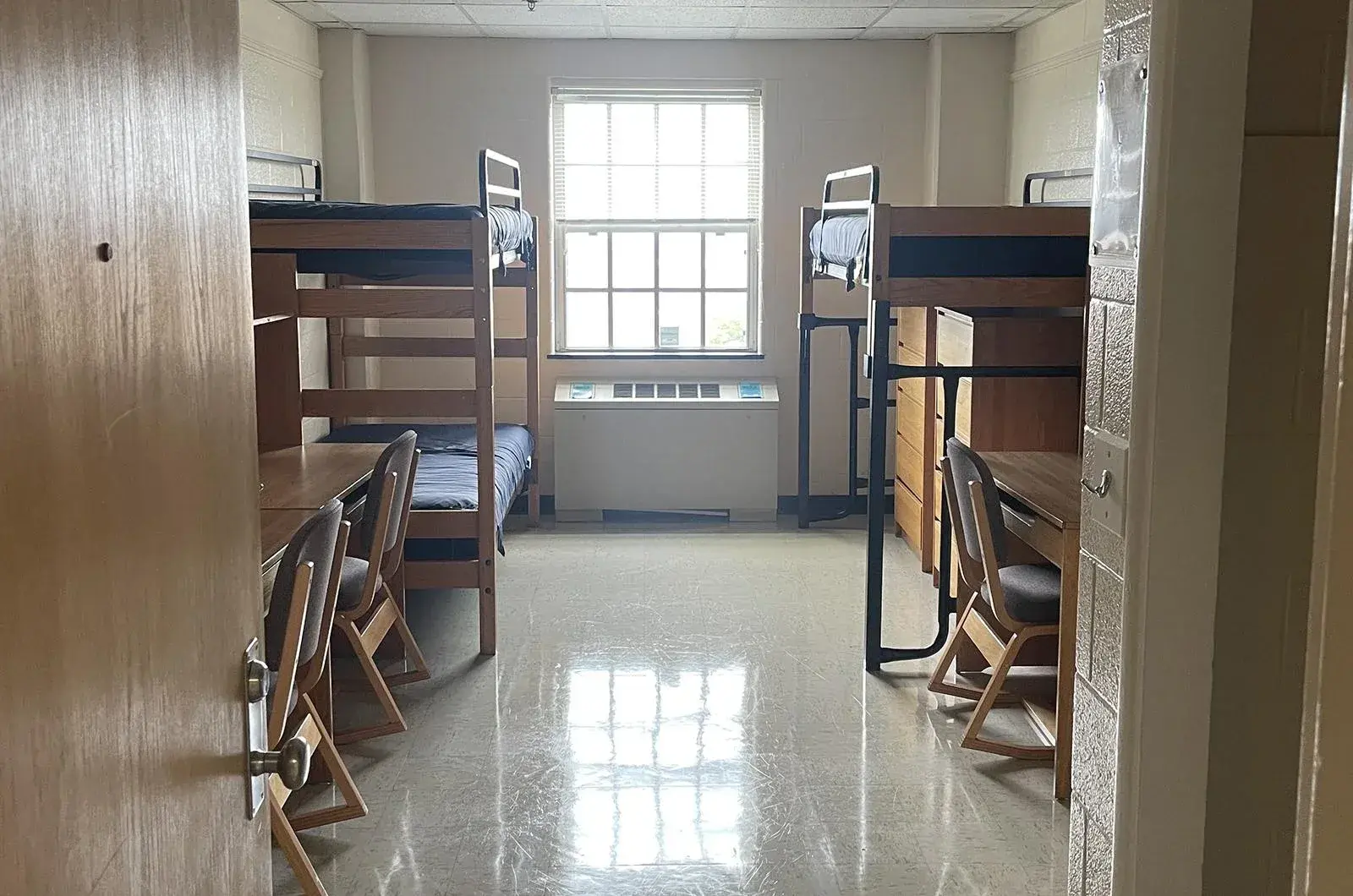 Empty dorm room with a set of bunk beds and a single with 2 dressers underneath bed. @ desks with chairs.