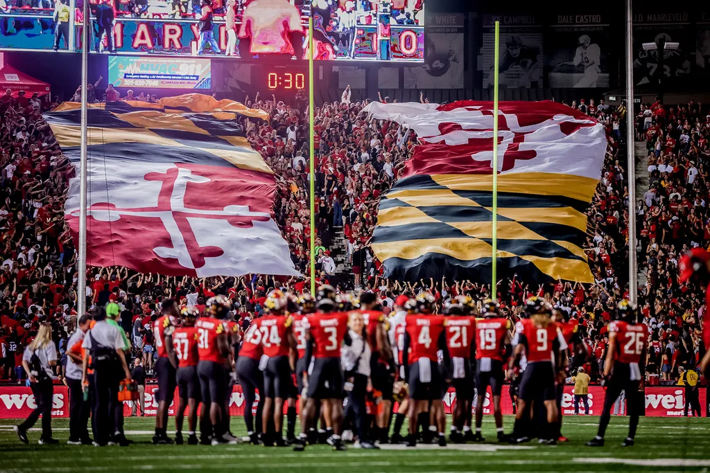 Football players gathered on field. In stands, huge flag of the state of Maryland.