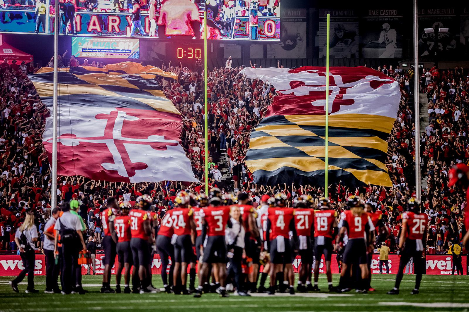 Football players gathered on field. In stands, huge flag of the state of Maryland.