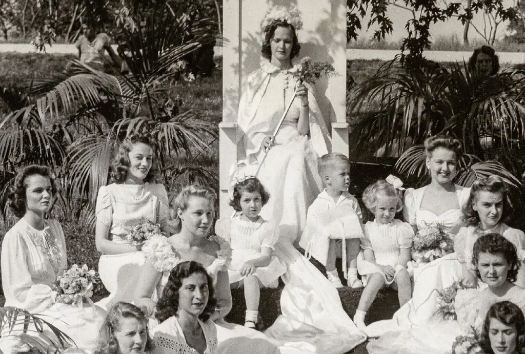 Vintage photo of young women in formal light dresses seated around a woman in a large seat. Women hold bouquets