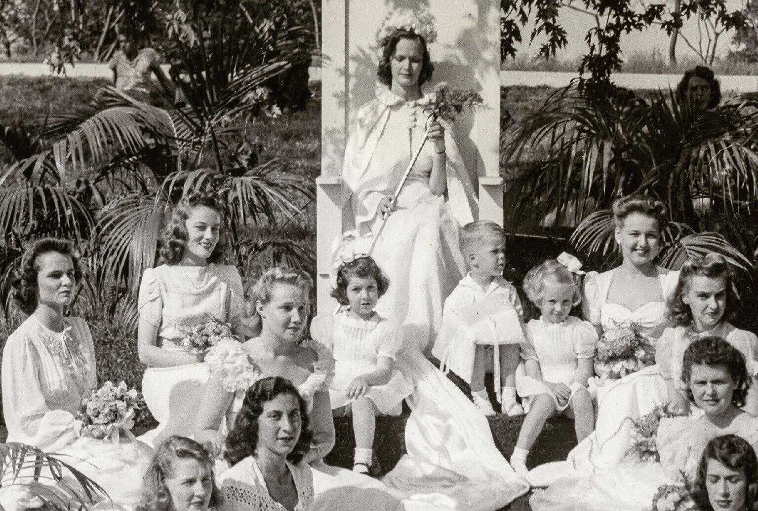 Vintage photo of young women in formal light dresses seated around a woman in a large seat. Women hold bouquets