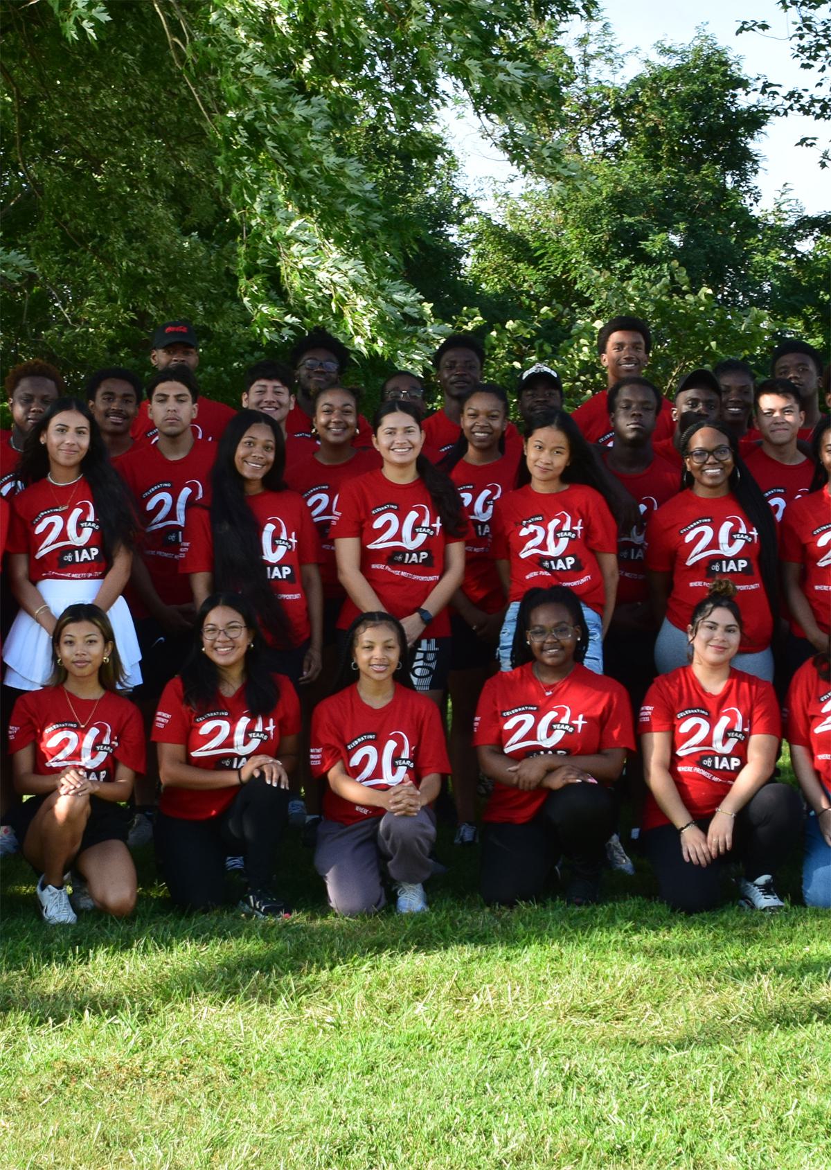 A group of young people gathered outside on a green lawn, each of them wearing a red t-shirt with white numbers "20+"