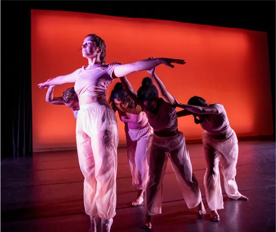 Group of performers on stage mid-pose one dancer holds their arms out stretched, the others from behind hold them up