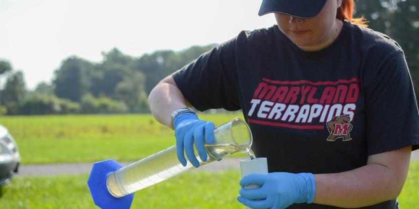 A student wearing nylon gloves pours liquid from a tall beaker into a container with an open field and trees surrounding in the background.