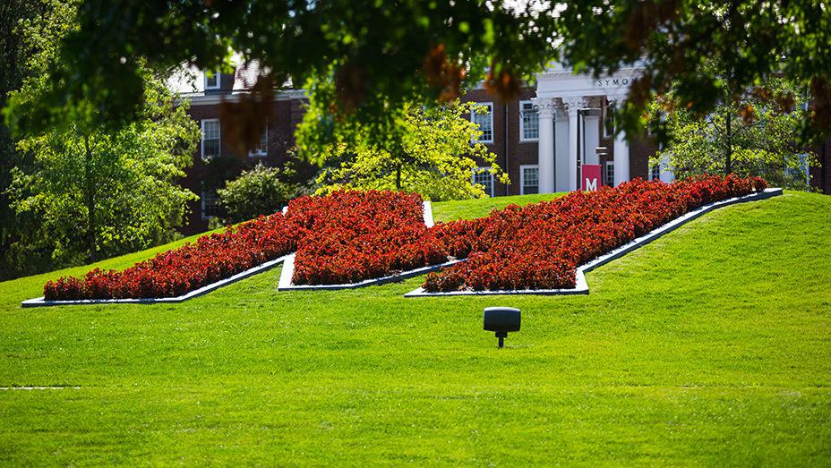 large "M" in flowers on a lawn in front of a traditional building