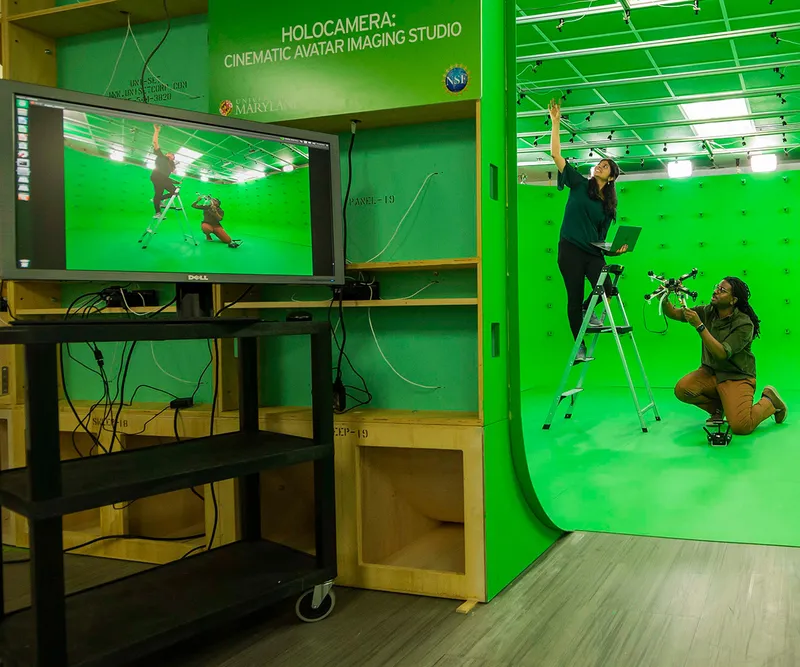 2 people working in a green room with AV equipment. Sign says "Holocamera: Cinematic Avatar Imaging Studio"