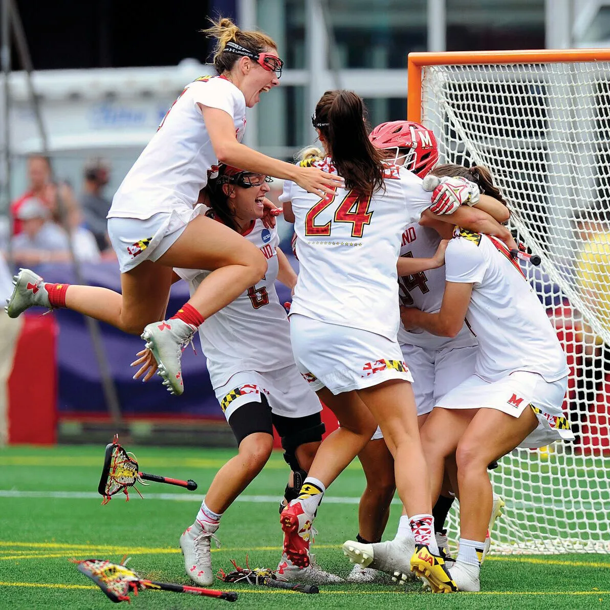 Excited women's lacrosse players in front of the net. One is jumping in the air.
